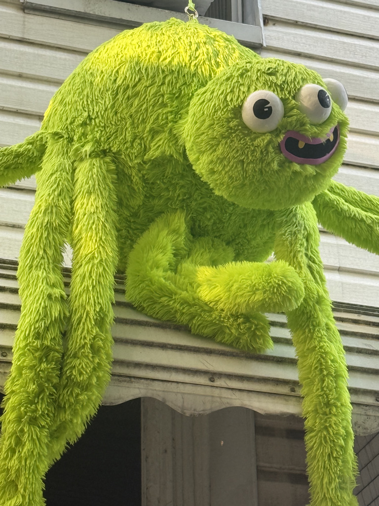 A large, fluffy, bright green plush creature with big eyes and a playful expression, hanging from the edge of a building.
